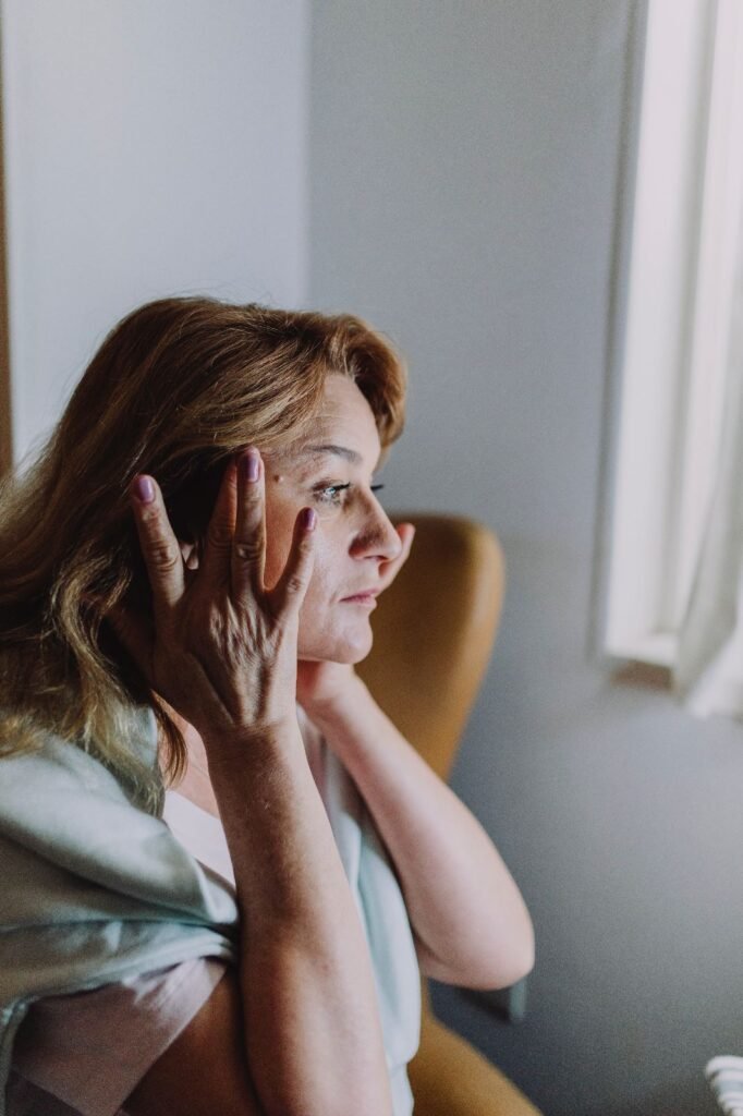 Middle-aged woman with light brown hair touching her temples while looking into the distance, sitting near a window with soft natural light